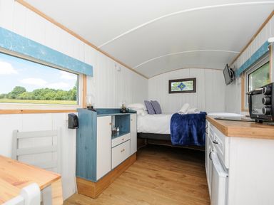 A small room with a bed at the end a blue and white cabinet a wooden table and chair and kitchen appliances at Shepherds Hut Boconnoc near Lostwithiel