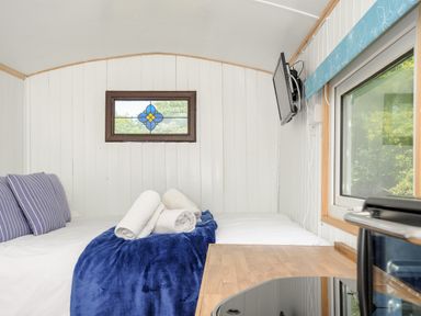 A small room with a bed with pillows and rolled towels a wall-mounted television and a window at Shepherds Hut in Boconnoc near Lostwithiel