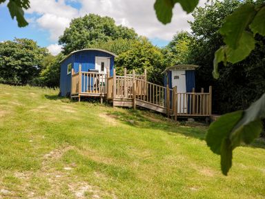 A blue shepherds hut with a wooden deck and ramp and a small blue bathing hut surrounded by trees at Shepherds Hut in Boconnoc near Lostwithiel