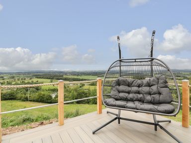 An outdoor deck with a hanging cushioned swing chair overlooking fields and hills at Laird Lodge 5 in Lochmaben
