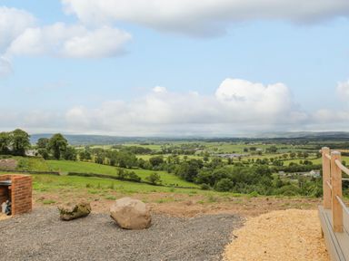 A rural landscape with green fields trees and distant buildings seen from a gravel area near a wooden deck with railing at Laird Lodge 5 in Lochmaben