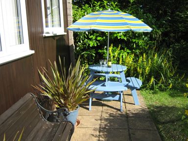 An outdoor patio with a wooden bench a blue round table with an umbrella and plants at 8 The Park in Kilkhampton