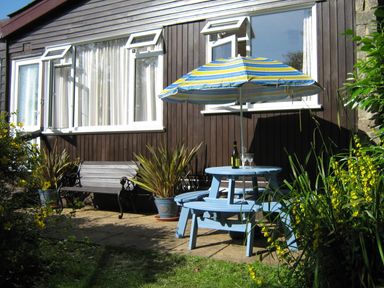 An outdoor patio with a wooden bench two potted plants a blue round picnic table with a striped umbrella and a bottle with glasses at 8 The Park in Kilkhampton