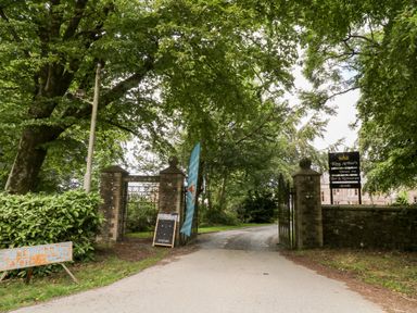 An entrance gate with stone pillars and metal gates surrounded by trees and signs for King Arthurs venue and a swimming pool at 8 The Park in Kilkhampton