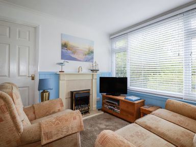 A living room with armchairs a fireplace a television on a wooden stand and a large window with blinds at 8 The Park in Kilkhampton