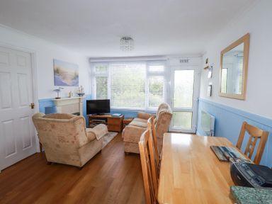 A living room with beige armchairs a wooden dining table a television by the window and a door at 8 The Park in Kilkhampton