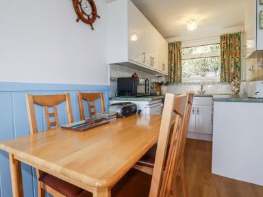 A kitchen with a wooden dining table and chairs a microwave and a window with patterned curtains at 8 The Park in Kilkhampton