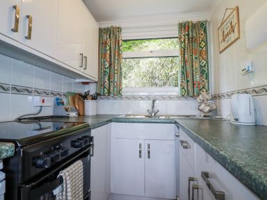 A kitchen with a stove, sink, kettle, cabinets, utensil holder, patterned curtains, and a wall sign at 8 The Park in Kilkhampton