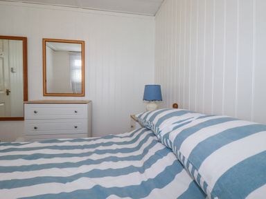 A bedroom with a bed with blue and white striped bedding a dresser with two mirrors and a bedside table with a blue lamp at 8 The Park in Kilkhampton