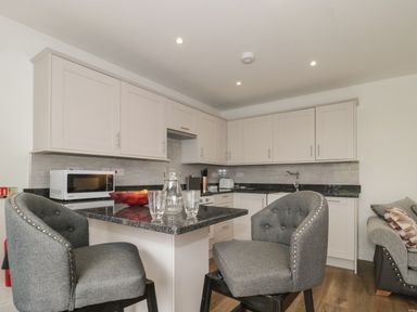 A kitchen with white cabinets a microwave on the counter a black countertop with chairs and glassware at The Hideaway in Lympsham near Brean