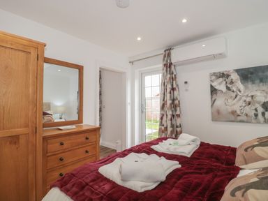 A bedroom with a bed covered in a red blanket and white towels on it a wooden dresser with a mirror a door leading outside curtains and a wall-mounted air conditioner at The Hideaway in Lympsham near Brean