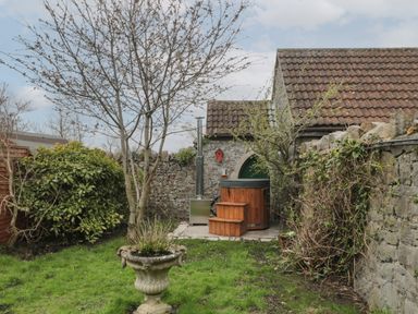 A garden with a tree and a wooden hot tub near stone walls and a tiled roof building at The Hideaway in Lympsham near Brean