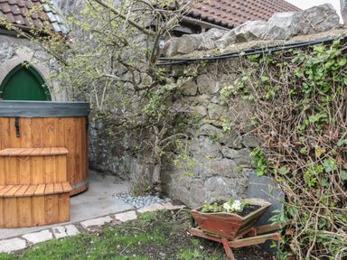 An outdoor space with a wooden hot tub next to a stone wall and a wheelbarrow planter with flowers at The Hideaway in Lympsham near Brean