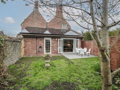 A backyard with a grassy lawn a tree a stone wall and a patio with white metal chairs and a table at The Hideaway in Lympsham near Brean