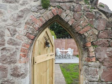 An arched stone doorway with a wooden gate opening to a patio with white metal table and chairs at The Hideaway in Lympsham near Brean