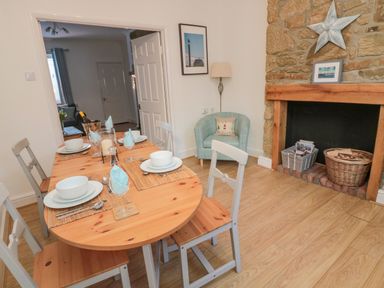 A dining room with a wooden table set for four next to a stone fireplace with baskets and a blue armchair at Redshank in Amble