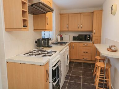 A kitchen with wooden cabinets a gas stove a washing machine a microwave toaster and kettle with a small breakfast bar and stools at Redshank in Amble