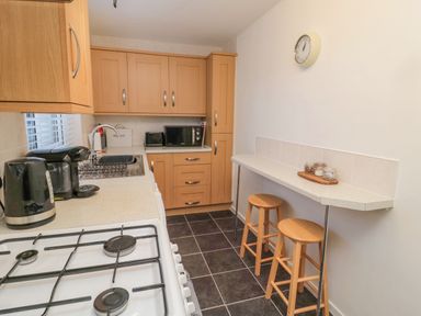 A kitchen with wooden cabinets a white gas stove black appliances two wooden stools and a wall clock at Redshank in Amble