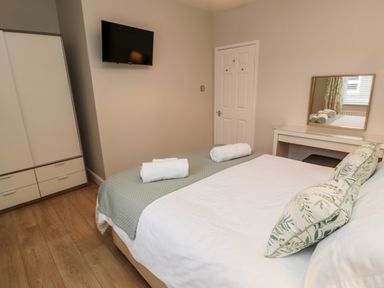 A bedroom with a bed with pillows and rolled towels a wardrobe a wall-mounted tv and a dressing table with a mirror at Redshank in Amble