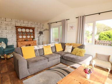 A living room with a grey sofa with yellow cushions a wooden cabinet displaying plates and a wooden coffee table with flowers at Dragonfly Lodge in Worthen near Minsterley