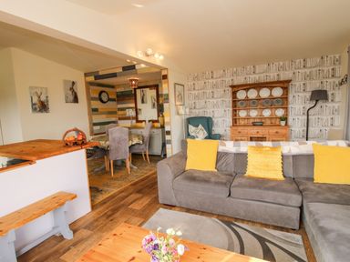 A living room with a gray sofa with yellow cushions a wooden coffee table a dining area with a table and chairs and a wooden cabinet with plates at Dragonfly Lodge in Worthen near Minsterley