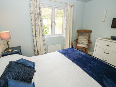 A bedroom with a bed and pillows a wooden chair with cushions a white dresser and a window with floral curtains at Dragonfly Lodge in Worthen near Minsterley