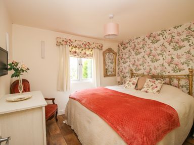 A bedroom with a bed with floral pillows and a red blanket a floral wallpaper a window with curtains a mirror a chair and a TV on the wall at Dragonfly Lodge in Worthen near Minsterley