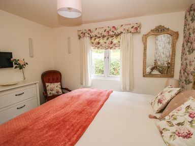 A bedroom with a bed covered by a coral blanket and floral pillows a red chair floral curtains and a gold framed mirror at Dragonfly Lodge in Worthen near Minsterley