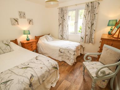 A bedroom with two single beds a wooden nightstand with a lamp a wooden dresser a window with curtains and an upholstered chair at Dragonfly Lodge in Worthen near Minsterley
