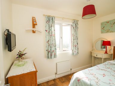 A bedroom with a bed covered in floral bedding a wooden dresser with a vase of flowers a mounted television and a dressing table with a red lamp at Dragonfly Lodge in Worthen near Minsterley