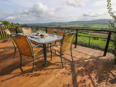 An outdoor deck with a glass table and six chairs overlooking a rural landscape at Dragonfly Lodge in Worthen near Minsterley