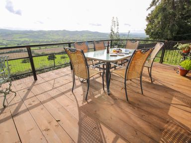 An outdoor wooden deck with a glass top dining table and six wicker chairs overlooking green fields at Dragonfly Lodge in Worthen near Minsterley