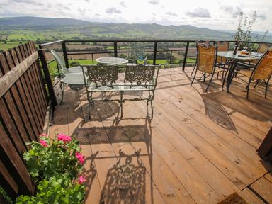 An outdoor wooden deck with metal chairs and tables overlooking a valley and hills at Dragonfly Lodge in Worthen near Minsterley