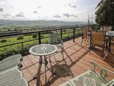 A wooden deck with metal and wicker outdoor furniture overlooking green fields and hills at Dragonfly Lodge Worthen near Minsterley