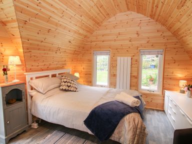 A bedroom with a double bed bedside tables lamps and dresser in a wooden paneled room with two windows at The Pod Worthen near Minsterley