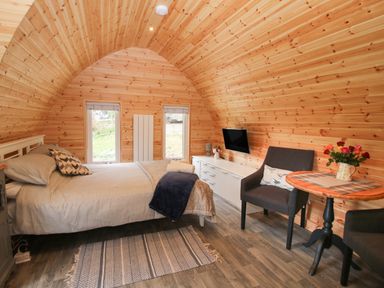A bedroom with wooden walls a bed two windows a dresser a mounted television two chairs and a small round table at The Pod in Worthen near Minsterley