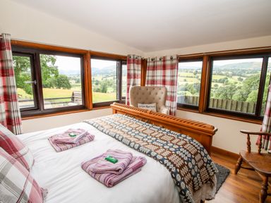 A bedroom with a wooden bed frame an armchair and windows showing a countryside view at The Retreat in Pedairffordd near Llanrhaeadr-Ym-Mochnant