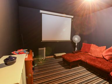 A room with a red couch and ottoman striped carpet a white fan a heater and a projector screen at The Retreat in Pedairffordd near Llanrhaeadr-Ym-Mochnant