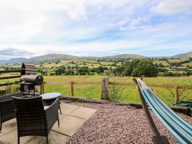 An outdoor patio area with wicker chairs and a table a barbecue grill and a blue hammock overlooking a green field with hills at The Retreat in Pedairffordd near Llanrhaeadr-Ym-Mochnant