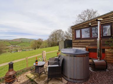 An outdoor seating area with a hot tub two chairs a small table with drinks and a fireplace near a wooden cabin at The Retreat in Pedairffordd near Llanrhaeadr-Ym-Mochnant