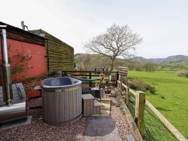 An outdoor area with a wooden hot tub, chairs, a hammock, a small table with drinks, and a chiminea overlooking green fields at The Retreat in Pedairffordd near Llanrhaeadr-Ym-Mochnant