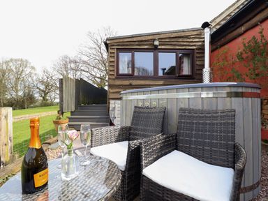 An outdoor seating area with two wicker chairs and a glass table with a bottle and glasses next to a wooden hot tub at The Retreat in Pedairffordd near Llanrhaeadr-Ym-Mochnant