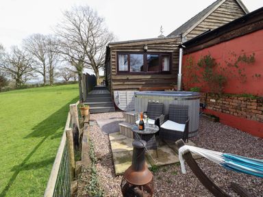 An outdoor area with a hot tub two chairs a small table with a bottle and glasses a firepit and a hammock next to a wooden building at The Retreat in Pedairffordd near Llanrhaeadr Ym Mochnant