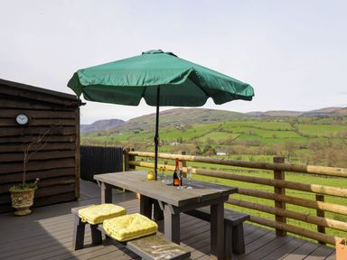 An outdoor deck with a wooden picnic table and benches with yellow cushions under a green umbrella overlooking green hills at The Retreat in Pedairffordd near Llanrhaeadr-Ym-Mochnant