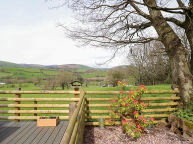 A wooden fenced patio with potted plant and a large tree with a green field and hills in the background at The Retreat in Pedairffordd near Llanrhaeadr-Ym-Mochnant