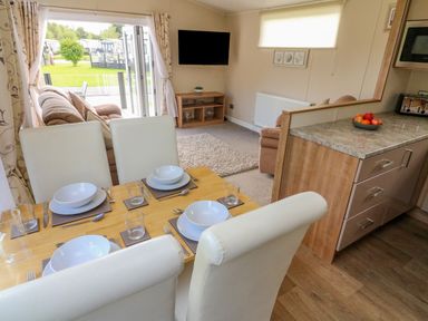 A dining area with a wooden table set for four with white chairs next to a living room with sofas a wall-mounted TV and an open door leading to a garden at Number 15 Carnforth