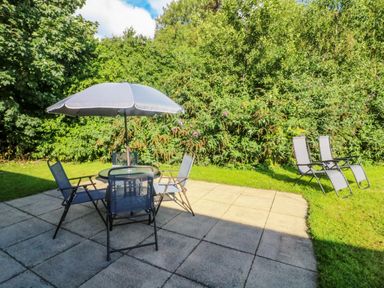 A patio area with a round table with an umbrella and four chairs and two reclining chairs on a lawn surrounded by trees and bushes at Number 15 in Carnforth