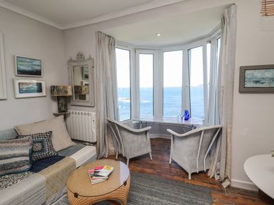 A living room with a sofa pillows round table two wicker chairs and windows overlooking the sea at Starfish in Sennen Cove