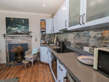 A kitchen with white cabinets a countertop a sink a microwave a stove and a chair near a fireplace with a painting above at Starfish in Sennen Cove