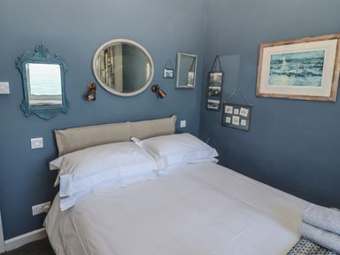 A bedroom with a bed with white linens and pillows and multiple mirrors and framed pictures on blue walls at Starfish in Sennen Cove